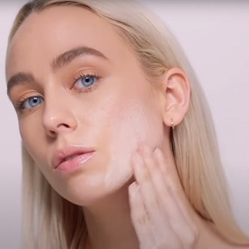 A young woman with blue eyes applies a gentle cleanser to her cheek for a fresh, glowing look.