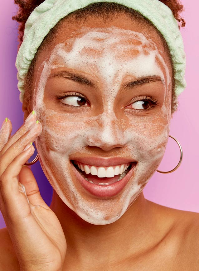 Woman cleansing her face with a foamy wash, smiling against a pink background, showcasing a deep-cleaning skincare routine.