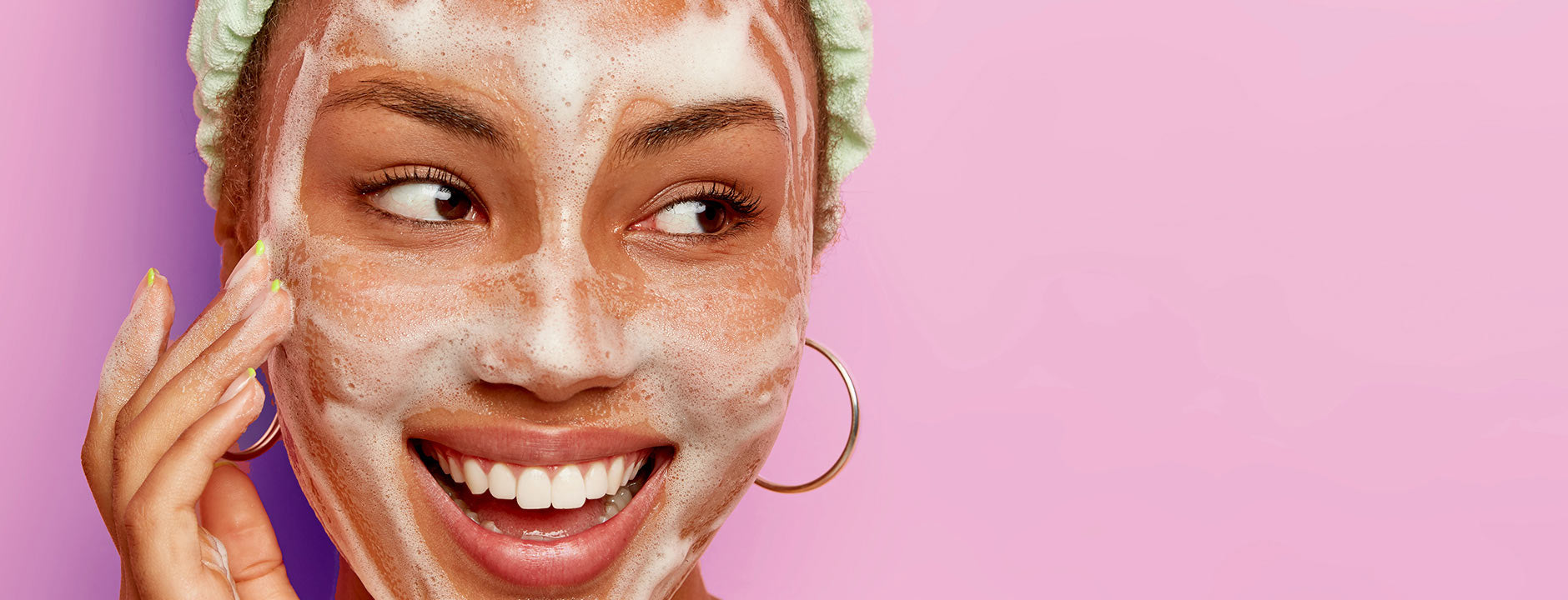 Woman cleansing her face with a foamy wash, smiling against a pink background, showcasing a deep-cleaning skincare routine.