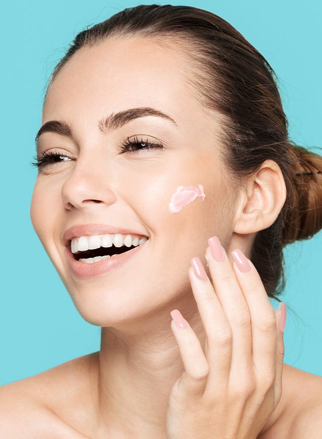 Smiling woman applying pink cream to her cheek against a bright blue background, promoting skincare.