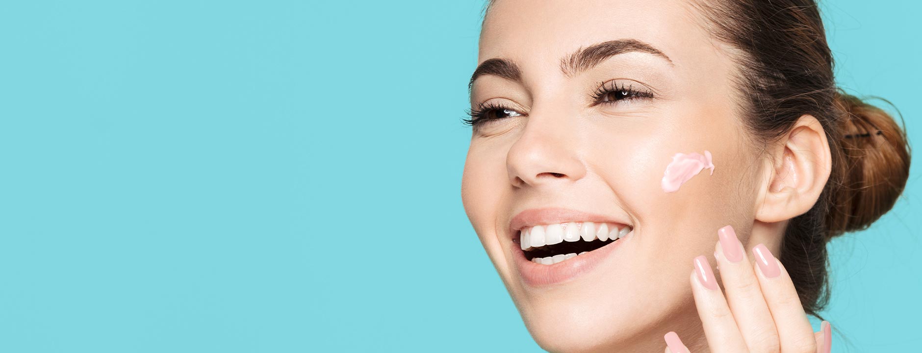 Smiling woman applying pink cream to her cheek against a bright blue background, promoting skincare.